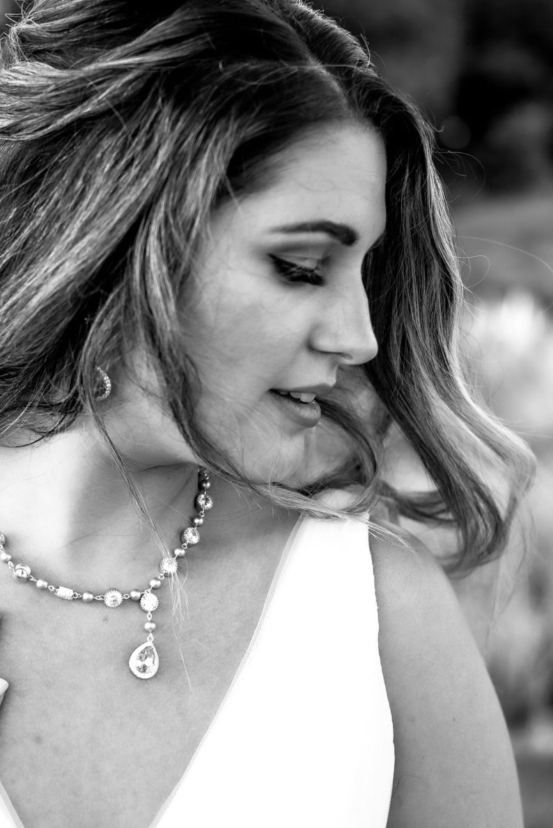 May include: A black and white portrait of a person wearing a sparkling necklace with a teardrop pendant. The necklace is made of small, round beads and larger, clear stones. The person is wearing a white dress or top.