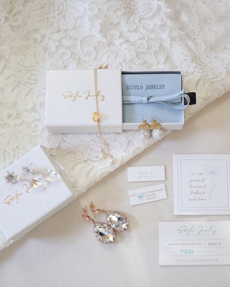 May include: An assortment of jewelry pieces, including earrings and a necklace, presented in white boxes with gold lettering. The image also features business cards and small tags, all arranged on a white lace surface.