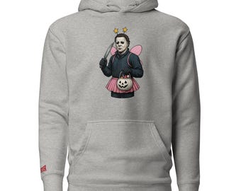 Puede incluir: Una sudadera con capucha gris claro con una ilustración humorística de Michael Myers con un disfraz de hada. El gráfico muestra a Myers sosteniendo un cuchillo, usando un tutú rosa, alas de hada y una bolsa de golosinas de calabaza. La sudadera tiene un bolsillo de canguro y una capucha con cordón.
