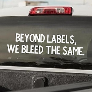 May include: A white decal on a vehicle's rear window. The decal reads "BEYOND LABELS, WE BLEED THE SAME" in a sans-serif font. The vehicle is a light-colored pickup truck, with a black bed.