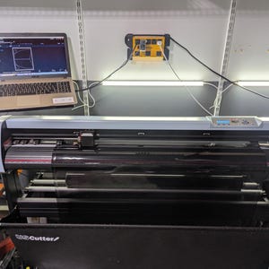 May include: A silver and black vinyl cutter with a black stand. The cutter has a clear protective cover over the cutting blade. A laptop computer is connected to the cutter and shows a design on the screen.