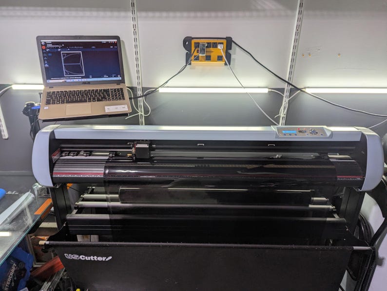 May include: A silver and black vinyl cutter with a black stand. The cutter has a clear protective cover over the cutting blade. A laptop computer is connected to the cutter and shows a design on the screen.