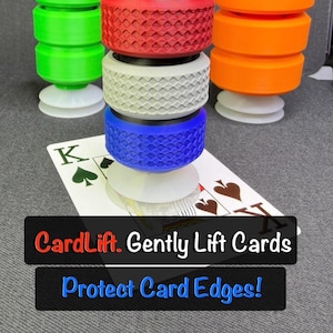 May include: Three cylindrical card lifters with suction cups, one green, one orange, and one stacked with red, white, and blue textured segments. A playing card is underneath the center lifter. Text reads: CardLift. Gently Lift Cards Protect Card Edges!
