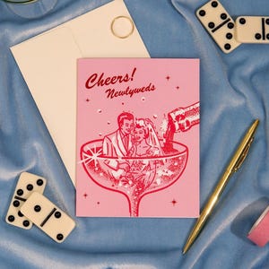 May include: A pink wedding card with the words "Cheers! Newlyweds" and an illustration of a couple in a champagne glass. The card is next to a white envelope, a gold pen, and dominoes on a blue fabric.