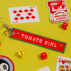 May include: A selection of novelty items on a yellow background. Includes a red keychain with "TOMATO GIRL" text, playing cards, dice, and a patch that says "HANDING MY INNER CHILD A CIGARETTE".