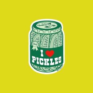 May include: A die-cut sticker featuring a green jar filled with pickles. The label displays the text I LOVE PICKLES with a red heart symbol. A small seal on the left reads Tasty. The jar lid shows a small cartoon pickle character illustration.