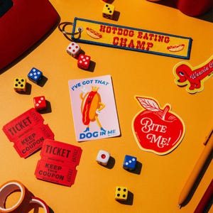 May include: A yellow fabric keychain with the text HOTDOG EATING CHAMP, a pink rectangular sticker with a cartoon hotdog and the text I'VE GOT THAT DOG IN ME, a red apple-shaped sticker with the text BITE ME, red tickets, and colourful dice.