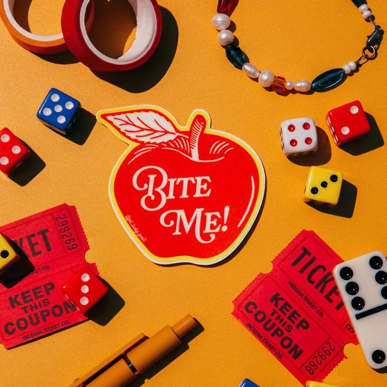 May include: A red apple-shaped sticker with the white text BITE ME! surrounded by red and white dice, two red raffle tickets, a beaded bracelet with white pearls and blue beads, two rolls of tape, and a yellow pen on a solid orange background.