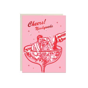 May include: A pink wedding card featuring a red illustration of a couple inside a champagne glass. The words "Cheers! Newlyweds" are written in red script above the couple. A bottle pours champagne.