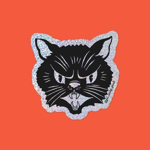 May include: A black cat sticker with an angry expression, white fangs, and whiskers. The sticker has a silver glitter border and is set against an orange background. The text "@get_lucky_goods" is on the bottom right.