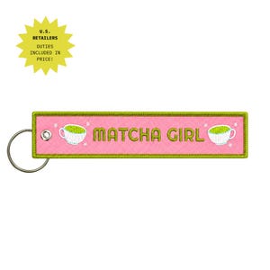 May include: A rectangular pink embroidered keyring with a lime green border and a silver metal ring. The text MATCHA GIRL is stitched in lime green, flanked by two white tea cup illustrations with green contents and small white star accents.