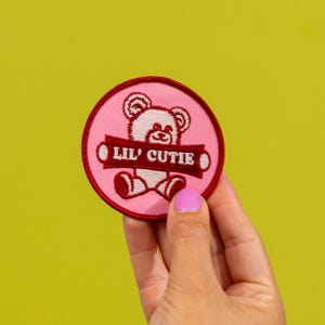 May include: A pink and red embroidered patch with a teddy bear design. The patch features the text "Lil' Cutie" in white letters on a red banner. The patch is round and has a pink background.