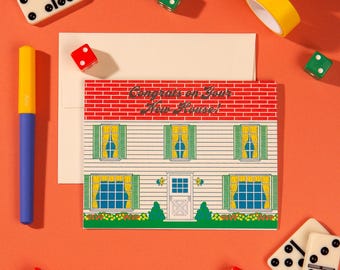 May include: A greeting card with a house illustration and the text "Congrats on Your New House!" is surrounded by dice, dominoes, and colorful tape rolls on an orange background. A yellow and blue pen is also present.