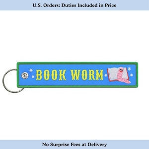 Peut inclure: Un porte-clés rectangulaire brodé bleu avec une bordure verte et un anneau en métal argenté. Le texte BOOK WORM est brodé en jaune, accompagné d'une illustration d'un livre ouvert et d'un ver rose portant des lunettes noires.