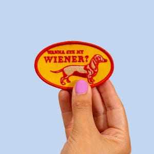 Peut inclure: Écusson brodé avec un fond ovale jaune et une bordure rouge. Le patch présente une illustration de chien teckel marron et un texte qui dit "WANNA SEE MY WIENER?" en rouge. Le patch est tenu par une main avec du vernis à ongles rose.