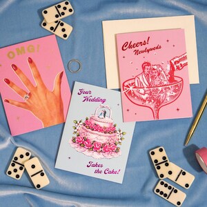May include: A selection of wedding cards and accessories on a blue surface. The cards feature illustrations of a hand with a ring, a wedding cake, and a couple in a champagne glass. Dominoes and a ring are also present.