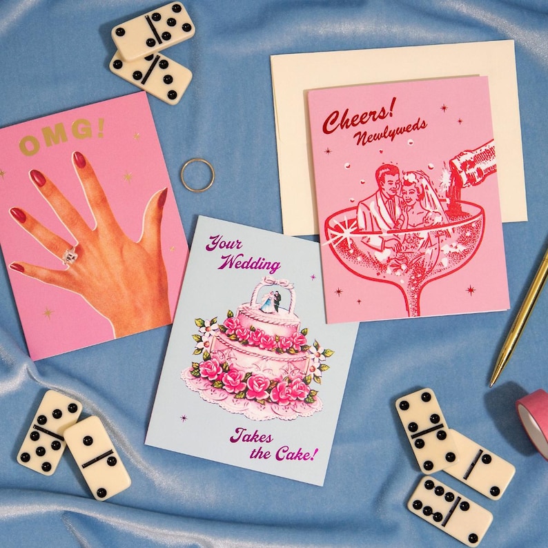 May include: A selection of wedding cards and accessories on a blue surface. The cards feature illustrations of a hand with a ring, a wedding cake, and a couple in a champagne glass. Dominoes and a ring are also present.