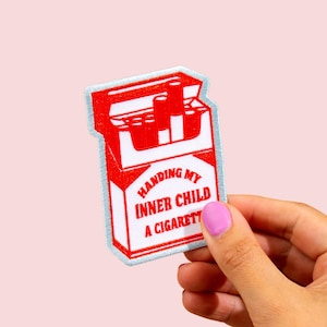 May include: Embroidered patch depicting a red and white cigarette packet with the text "HANDING MY INNER CHILD A CIGARETT". The patch has a light blue border. The background is a soft pink colour.