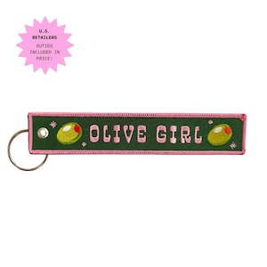 Peut inclure: Un porte-clés rectangulaire brodé vert foncé avec une bordure rose et un anneau en métal argenté. Le texte OLIVE GIRL est brodé en lettres roses, entouré de deux olives vertes avec des piments rouges et de petites étoiles roses.