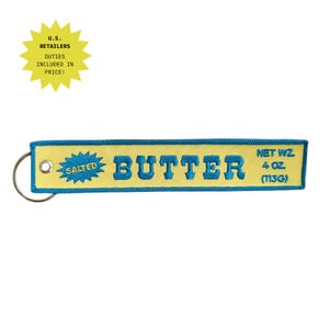 Peut inclure: Un porte-clés rectangulaire brodé jaune avec une bordure bleue et un texte bleu indiquant SALTED BUTTER et NET WT. 4 OZ. (113G). Le porte-clés comporte un anneau en métal argenté fixé à un œillet métallique sur le côté gauche de la bande.