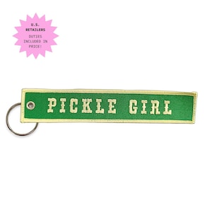May include: A rectangular green fabric keyring with a yellow border and the text PICKLE GIRL embroidered in yellow capital letters. The keyring features a silver metal grommet on the left side and a circular silver metal split ring attached.