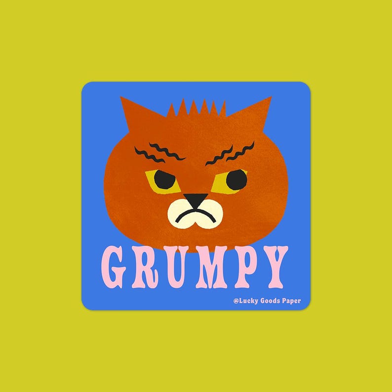 May include: A square sticker with a blue background featuring an orange cat head with a grumpy expression, yellow eyes, and wavy black eyebrows. The word GRUMPY is printed in large pink letters below the cat, with Lucky Goods Paper text.