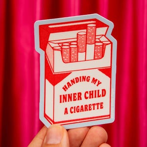 May include: A die-cut sticker featuring a red and white illustration of an open cigarette pack. The text on the front reads, "HANDING MY INNER CHILD A CIGARETTE." The design includes several cigarettes protruding from the top of the pack.