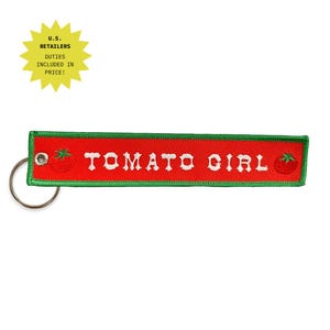 Peut inclure: Un porte-clés rectangulaire en tissu avec un fond rouge et une bordure verte. Le texte TOMATO GIRL est brodé en lettres blanches au centre. Deux petites illustrations de tomates rouges sont placées sur les côtés gauche et droit.