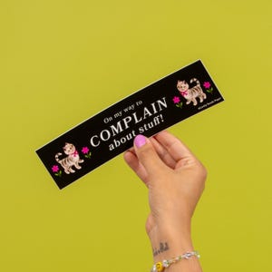 May include: A black sticker with the text "On my way to COMPLAIN about stuff!" features a cartoon cat and pink flowers. The sticker is held against a yellow-green background.