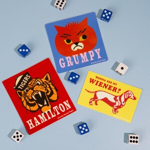 May include: Three rectangular stickers with graphic illustrations and six scattered dice. One sticker is blue with a red cat and the text GRUMPY. Another is red with a tiger and the text TIGERS HAMILTON. The third is yellow with a dachshund and the text WANNA SEE MY WIENER?