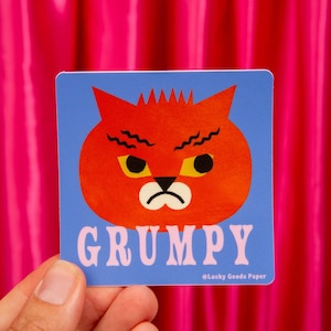 May include: A square sticker with a blue background featuring a stylized orange cat head with a grumpy expression, yellow eyes, and a black nose. The word GRUMPY is printed in large light pink letters below the cat, with Lucky Goods Paper text.