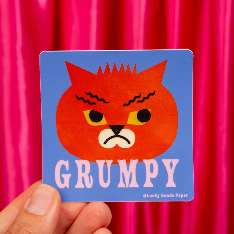 May include: A square sticker with a blue background featuring a stylized orange cat head with a grumpy expression, yellow eyes, and a black nose. The word GRUMPY is printed in large light pink letters below the cat, with Lucky Goods Paper text.