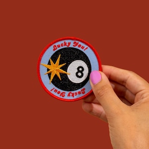Peut inclure: Un écusson rond avec un fond bleu clair et une bordure rouge. L'écusson présente une boule noire numéro 8, une étoile dorée et le texte "Lucky You!" en rouge.