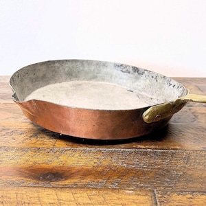 May include: A vintage copper frying pan with a brass handle. The pan has a worn, aged patina and is sitting on a wooden surface.