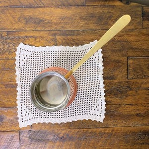 May include: A copper Turkish coffee pot with a long, thin, gold handle. The pot is sitting on a white crocheted doily.