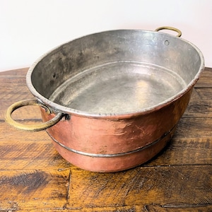 May include: Oval copper basin with brass handles. The basin has a hammered texture on the exterior and a smooth, silver-colored interior. The handles are attached to the rim of the basin. The copper has a reddish-brown hue.