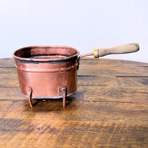 May include: A small copper pot with a wooden handle and three legs. The pot is oval shaped and has a dark patina.