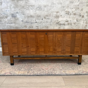 Puede incluir: Un baúl de almacenamiento rectangular de madera con un acabado de veta de madera marrón cálido y paneles verticales. El baúl se apoya sobre cuatro patas conectadas por una barra transversal horizontal e incluye un pequeño mecanismo de cierre.