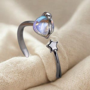 May include: A silver-toned ring with a large, iridescent, oval-shaped stone. The ring features a star-shaped accent on one end and a curved band that wraps around the finger. The stone has a blue and purple hue.
