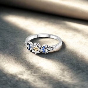 Op de afbeelding: Een zilveren ring met hemelse ontwerpen. De ring heeft een zon in het midden met gele accenten, geflankeerd door halve manen met blauwe stenen en sterren. De ring ligt op een grijze ondergrond.