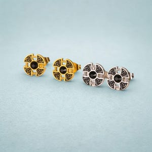 May include: Three pairs of stud earrings with a Celtic knot design. The earrings feature a circular shape with a black center stone. One pair is gold-toned, and the other is silver-toned, with black enamel detailing.