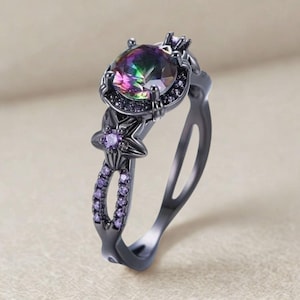 Peut inclure: Une bague noire ornée d'une grosse pierre irisée de forme ovale. La bague présente un motif en forme d'étoile avec des accents violets et de petites pierres violettes le long de l'anneau. Le métal de la bague a une finition sombre et mate.