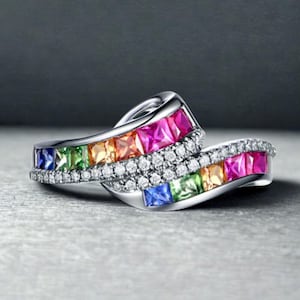 May include: A silver ring with a wave design, featuring a row of colourful gemstones in shades of blue, green, yellow, orange, and pink. The ring also has a row of small, clear stones. The ring is set against a grey background.