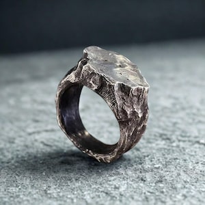 Op de afbeelding: Een zilverkleurige ring met een getextureerd, steenachtig uiterlijk. De ring heeft een onregelmatige, rechthoekige bovenkant en een dikke band, met een ruw, organisch ontwerp. Het oppervlak heeft een ruwe, ongelijke textuur.