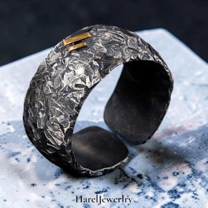 Könnte beinhalten: Ein breiter Ring mit offenem Band in dunkler, oxidierter Silberoptik mit einer stark texturierten, zerklüfteten Oberfläche. Ein kleiner, rechteckiger goldfarbener Akzent ist nahe der Oberkante eingelassen. Der Text HarelJewelry steht unten.