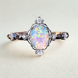 Op de afbeelding: Zilverkleurige ring met een ovale opaal edelsteen met een regenboogkleurige iriserende glans. De ring is versierd met kleine, heldere edelstenen en decoratieve details op de band. Een sieraad.