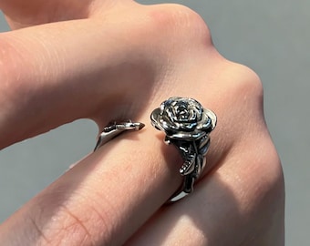 Könnte beinhalten: Ein silberfarbener Ring mit einem detaillierten Rosen-Design. Der Ring umschließt den Finger, wobei die Rosenblüte oben liegt. Das Design umfasst Blätter und Stiele und erzeugt eine realistische florale Ästhetik.