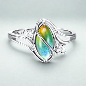 Può includere: Un anello d'argento con una pietra centrale ovale cangiante, che mostra sfumature di giallo, verde e blu. Due pietre di accento chiare e rotonde affiancano la pietra centrale. L'anello è contrassegnato con "S925".