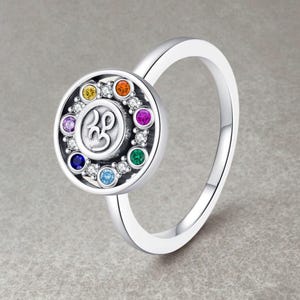 Può includere: Anello d'argento con un pendente circolare con un design centrale e un anello di pietre preziose colorate. L'anello ha una finitura lucida e una fascia semplice. Le pietre preziose sono in vari colori, tra cui blu, verde e arancione.