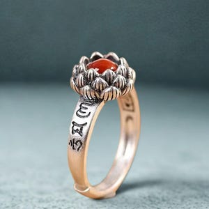 May include: A silver-coloured ring with a lotus flower design and a red gemstone. The band has etched symbols. The ring is set against a teal background.